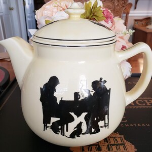 May include: Cream-colored ceramic teapot with a black silhouette design of two people seated at a table with a dog. The teapot has a rounded shape, a lid, and a curved handle. Black trim accents the lid.
