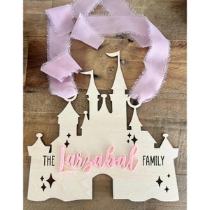 Könnte beinhalten: Ein Holzausschnitt eines Disney-Schlosses mit einem rosa Band, das oben hängt. Das Schloss hat ein rosa Schild mit der Aufschrift "The Larzabal Family".