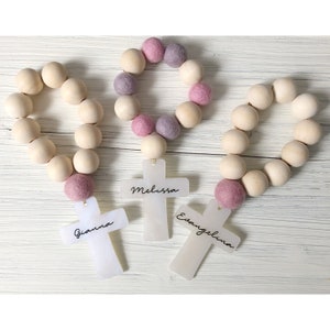 May include: Three wooden bead rosaries with pink and purple felt balls. Each rosary has a white cross with a name on it: Gianna, Melissa, and Evangelina.