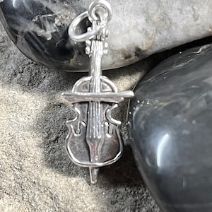 May include: A silver-colored charm in the shape of a cello, featuring detailed strings, a bridge, and a scroll. The charm has a loop at the top for attachment. The cello charm is displayed against a backdrop of gray and black stones.