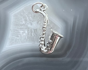 May include: A silver saxophone charm pendant. The charm is detailed with a textured surface and a loop for attaching to a chain or bracelet.