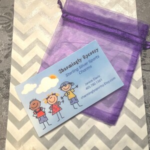 May include: A white paper bag with a chevron pattern and a purple organza bag. The bag has a business card that says "Charmingly Sporty Sterling Silver Sports Charms" with a phone number and website address. The business card has a cartoon illustration of three children.