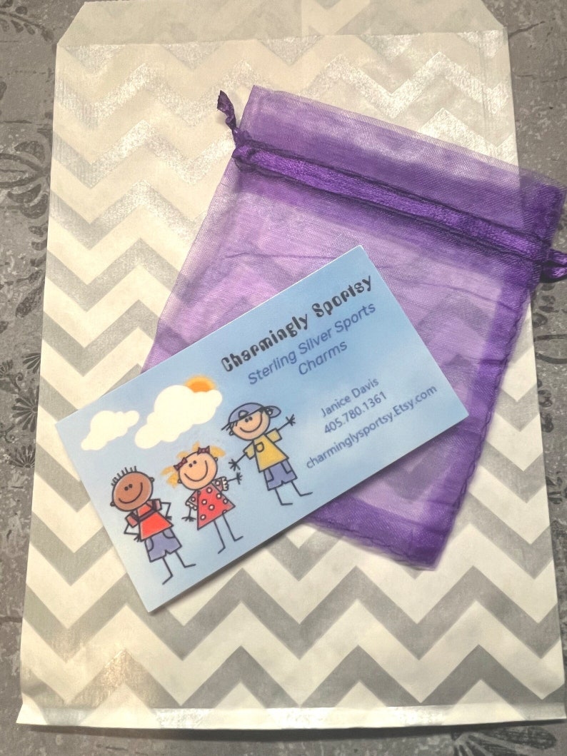 May include: A white paper bag with a chevron pattern and a purple organza bag. The bag has a business card that says "Charmingly Sporty Sterling Silver Sports Charms" with a phone number and website address. The business card has a cartoon illustration of three children.