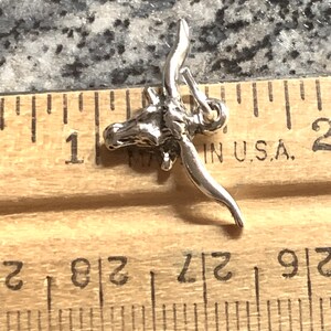 May include: Silver charm of a longhorn steer head with horns. The charm is about 1 inch long and is shown next to a ruler.