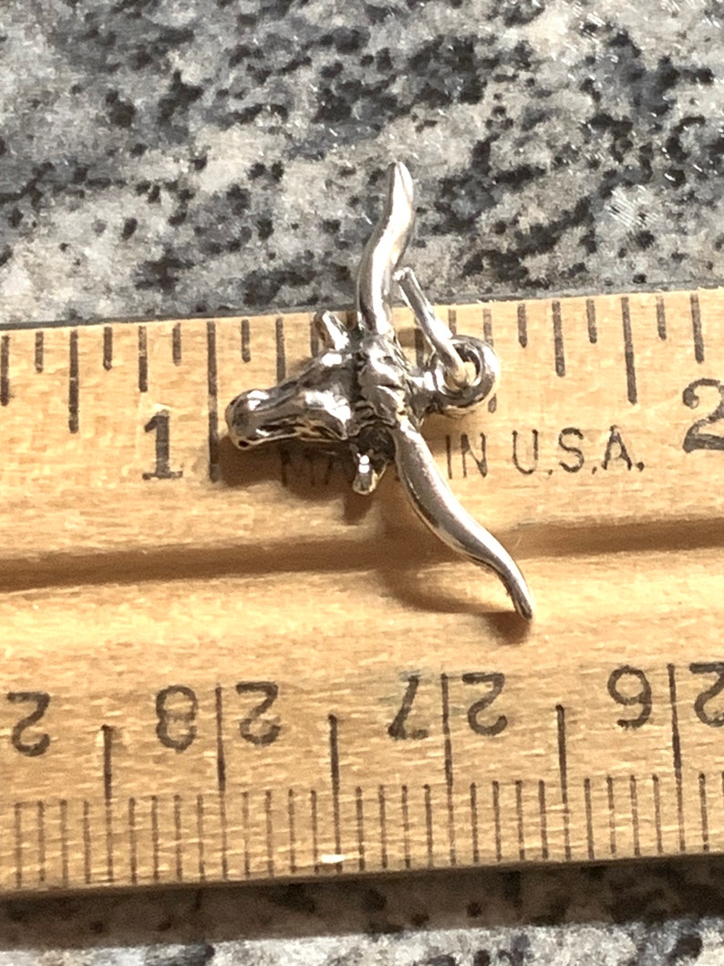 May include: Silver charm of a longhorn steer head with horns. The charm is about 1 inch long and is shown next to a ruler.