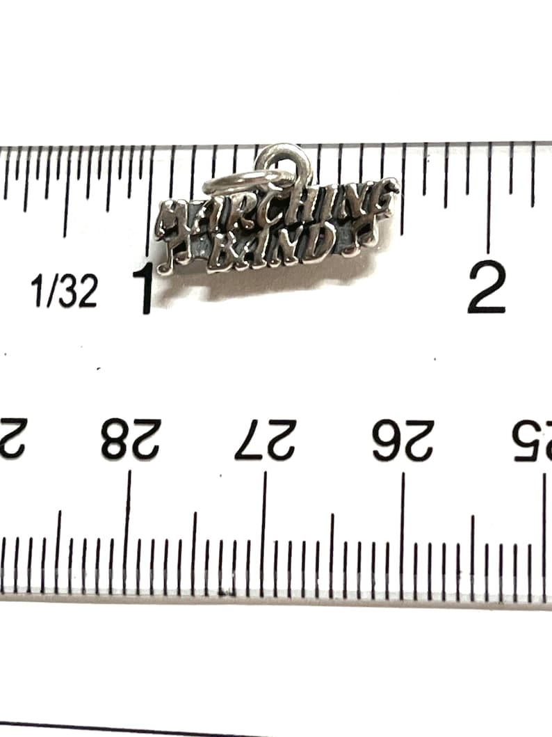 May include: Silver charm with the words "Marching Band" and a musical note. The charm is about 1 inch long and is measured against a ruler.
