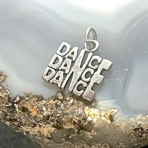 May include: Silver charm with the word "DANCE" repeated four times, forming a square shape. The charm has a loop at the top for attaching to a bracelet or necklace.