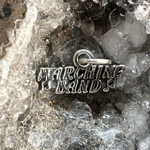 May include: A silver charm with the words "Marching Band" in a stylized font. The charm is sitting on a bed of white crystals.