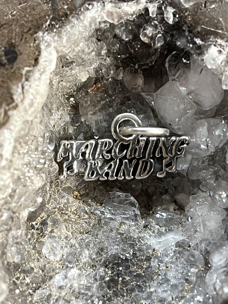 May include: A silver charm with the words "Marching Band" in a stylized font. The charm is sitting on a bed of white crystals.