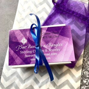 May include: A purple and white gift bag with a chevron pattern. A small white box with a purple and white design is inside the bag. The box has a blue ribbon tied around it. The box has the text "JSue Ban's n Bangles" and "Sterling Charms & Jewelry" printed on it. The text "Janice Davis" and a phone number are also printed on the box.