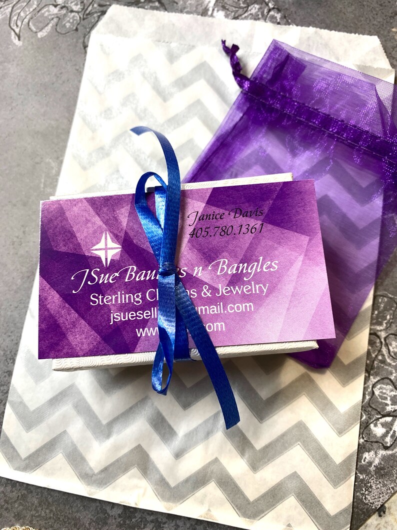 May include: A purple and white gift bag with a chevron pattern. A small white box with a purple and white design is inside the bag. The box has a blue ribbon tied around it. The box has the text "JSue Ban's n Bangles" and "Sterling Charms & Jewelry" printed on it. The text "Janice Davis" and a phone number are also printed on the box.