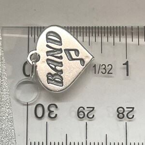 May include: Silver charm in the shape of a heart with the word "BAND" and a musical note engraved on it. The charm is about 1/2 inch wide.