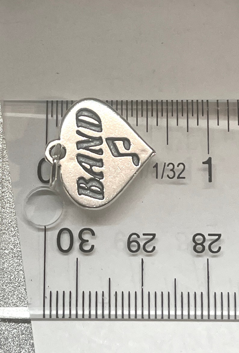 May include: Silver charm in the shape of a heart with the word "BAND" and a musical note engraved on it. The charm is about 1/2 inch wide.