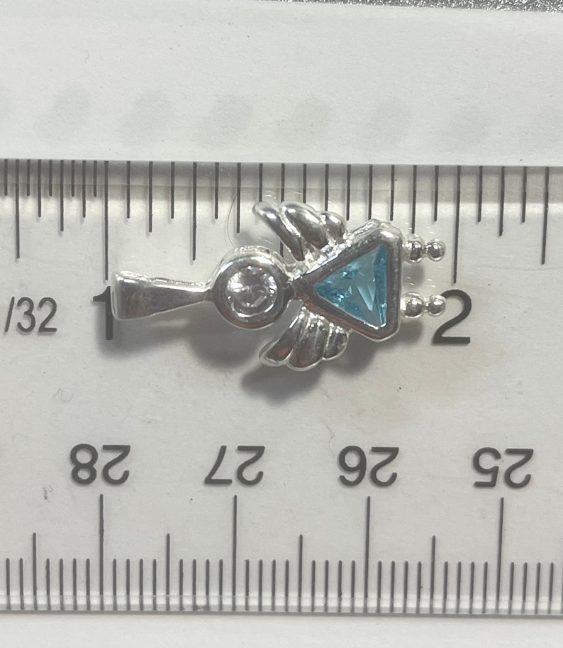 May include: Silver charm with a blue gemstone in the shape of a triangle, surrounded by wings and a small round gemstone. The charm is about 1 inch long.