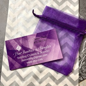 May include: A purple organza bag with a white and gray chevron patterned paper bag. The paper bag has a business card with the text "JSue Baubles n Bangles Sterling Charms & Jewelry jsuselling@gmail.com www.etsy.com" and the name "Janice Davis 405.780.1361".