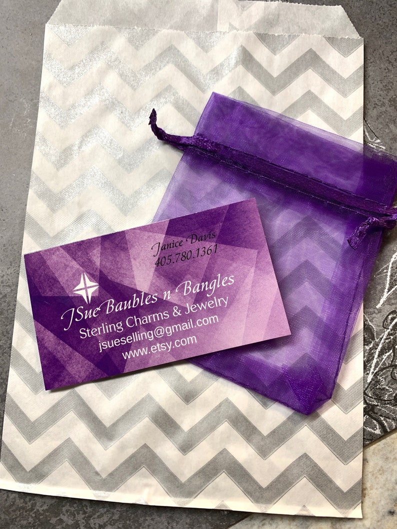 May include: A purple organza bag with a white and gray chevron patterned paper bag. The paper bag has a business card with the text "JSue Baubles n Bangles Sterling Charms & Jewelry jsuselling@gmail.com www.etsy.com" and the name "Janice Davis 405.780.1361".