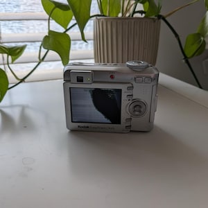 May include: A silver Kodak EasyShare C643 digital camera with a screen displaying a dark image. The camera is on a white surface, with a green plant and a beige pot in the background.