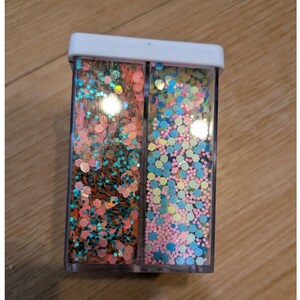 May include: A rectangular clear plastic container with a white lid, divided into two vertical compartments. The left side contains a mix of coral and teal hexagonal glitter, while the right side holds a blend of light blue, pink, and yellow glitter.