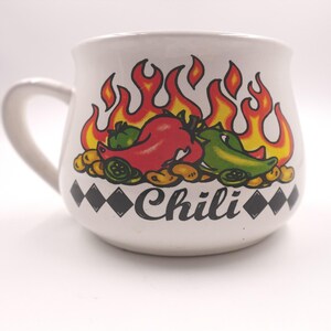 May include: A white ceramic mug with a handle featuring a colorful graphic of red and green chili peppers, beans, and orange flames. The word Chili is printed in black script below the graphic, flanked by a row of black diamond shapes on each side.