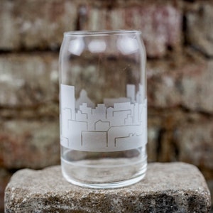 Peut inclure: Un verre à boire en verre transparent avec une ville gravée sur le côté. Le verre est posé sur une surface en pierre grise.