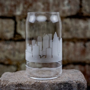 Peut inclure: Un verre en forme de canette transparent avec une silhouette de ville gravée sur le côté. La ville est blanche et le verre est transparent.