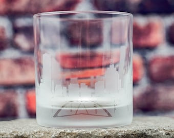 May include: A frosted glass tumbler with a cityscape etched into the side. The glass is sitting on a gray concrete block.