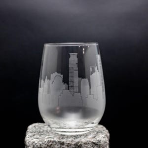 May include: A clear glass wine glass with an etched design of a city skyline. The glass is sitting on a gray stone surface.