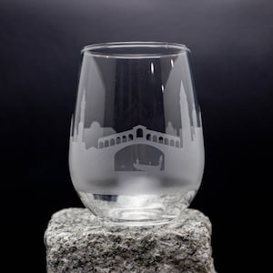 May include: A clear, stemless wine glass with an etched design of a cityscape, including a bridge and gondola. The glass is displayed on a gray stone against a black background. The glass is likely used for wine or other beverages.