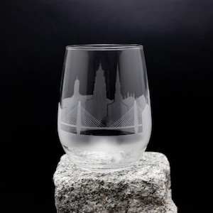 Peut inclure: Un verre à vin en verre transparent avec un motif gravé d'une ville et d'un pont. Le verre est posé sur un rocher gris.