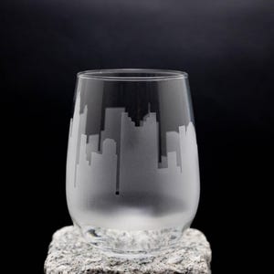 May include: A clear, stemless wine glass with a frosted skyline design. The cityscape silhouette wraps around the glass, creating a modern and elegant look. The glass is set on a textured, gray stone against a black background.