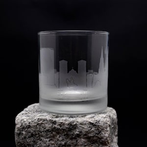 Puede incluir: Un vaso de vidrio esmerilado con un diseño de ciudad grabado. El vaso está sobre una roca gris.