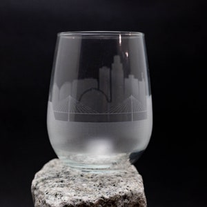 Puede incluir: Una copa de vino de vidrio transparente con un diseño esmerilado de un horizonte de ciudad y un puente. La copa está sobre una roca gris.