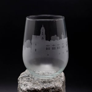 May include: A clear glass wine glass with an etched cityscape design. The glass is sitting on a gray stone.