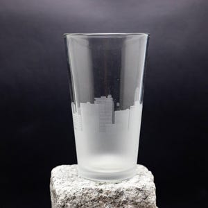 May include: A frosted glass pint glass with a cityscape etched into the side. The glass is sitting on a gray stone surface.