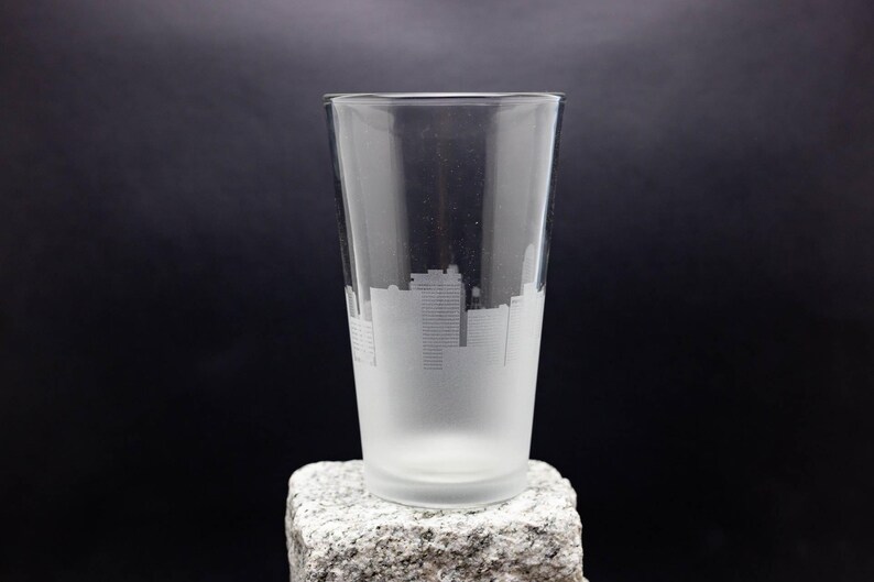 May include: A frosted glass pint glass with a cityscape etched into the side. The glass is sitting on a gray stone surface.