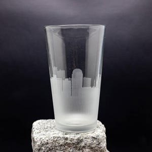 May include: A frosted glass pint glass with a cityscape etched into the side. The glass is sitting on a gray rock.