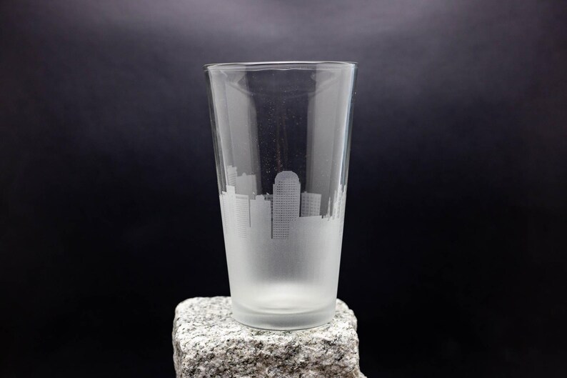May include: A frosted glass pint glass with a cityscape etched into the side. The glass is sitting on a gray rock.