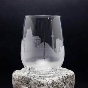 May include: A clear stemless wine glass with a city skyline etched into the glass. The glass is sitting on a gray stone against a black background. The etched design is white and features buildings and a mountain.