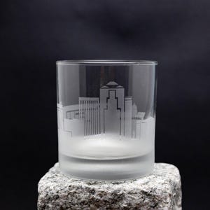 May include: A frosted glass tumbler with an etched cityscape design. The glass is sitting on a gray rock.