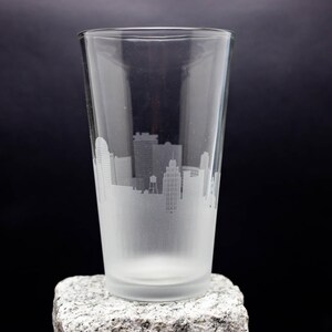 May include: A frosted glass pint glass with a cityscape etched into the side. The glass is sitting on a gray rock.