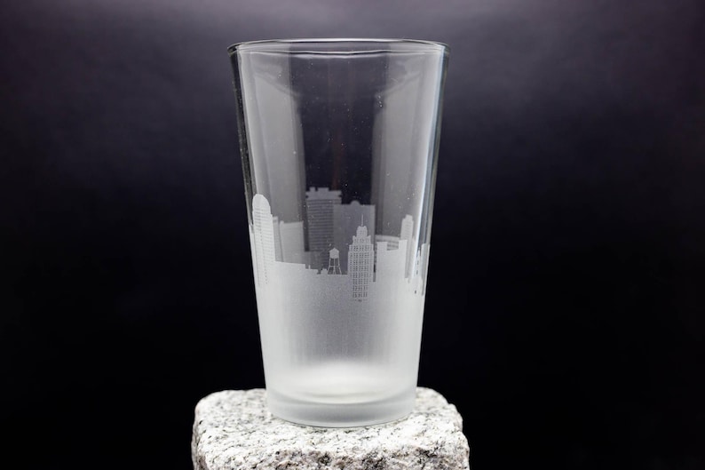 May include: A frosted glass pint glass with a cityscape etched into the side. The glass is sitting on a gray rock.