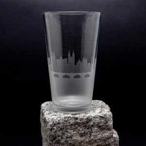 May include: A clear glass pint glass with a frosted design of a city skyline and bridge. The glass is sitting on a gray rock.