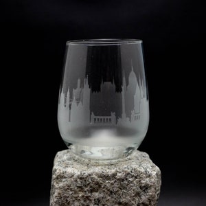 May include: A clear glass wine glass with an etched cityscape design. The glass is sitting on a gray stone.