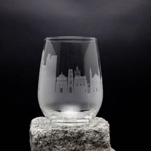 May include: A clear glass wine glass with a cityscape etched into the side. The glass is sitting on a gray rock.