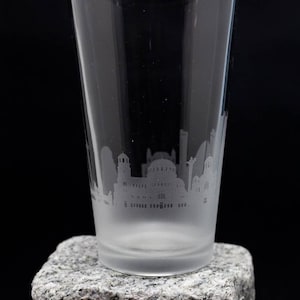 May include: A frosted glass pint glass with an etched cityscape design. The glass is sitting on a gray stone surface.