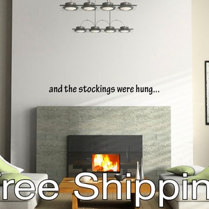 May include: A modern living room with a fireplace and the text "and the stockings were hung...". The fireplace has a stone facade and a burning fire. Two white sofas with green pillows flank a wooden coffee table.