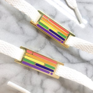 May include: Two white shoelaces with gold metal rainbow flag charms. The charms are enamel and feature a rainbow design with red, orange, yellow, green, blue, indigo, and violet stripes.