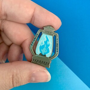 May include: A detailed enamel pin in the shape of a lantern. The pin features a light blue flame design inside a clear glass panel. The lantern has a metallic bronze finish with intricate detailing.
