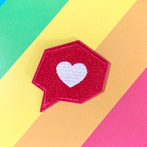 Puede incluir: Una burbuja de diálogo roja con un corazón blanco en el centro. La burbuja de diálogo es un parche de tela con un borde cosido.