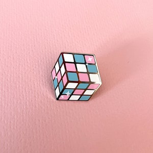 May include: A silver-coloured pin shaped like a Rubik's cube with pink, blue, and white squares. The pin has a sparkly finish.